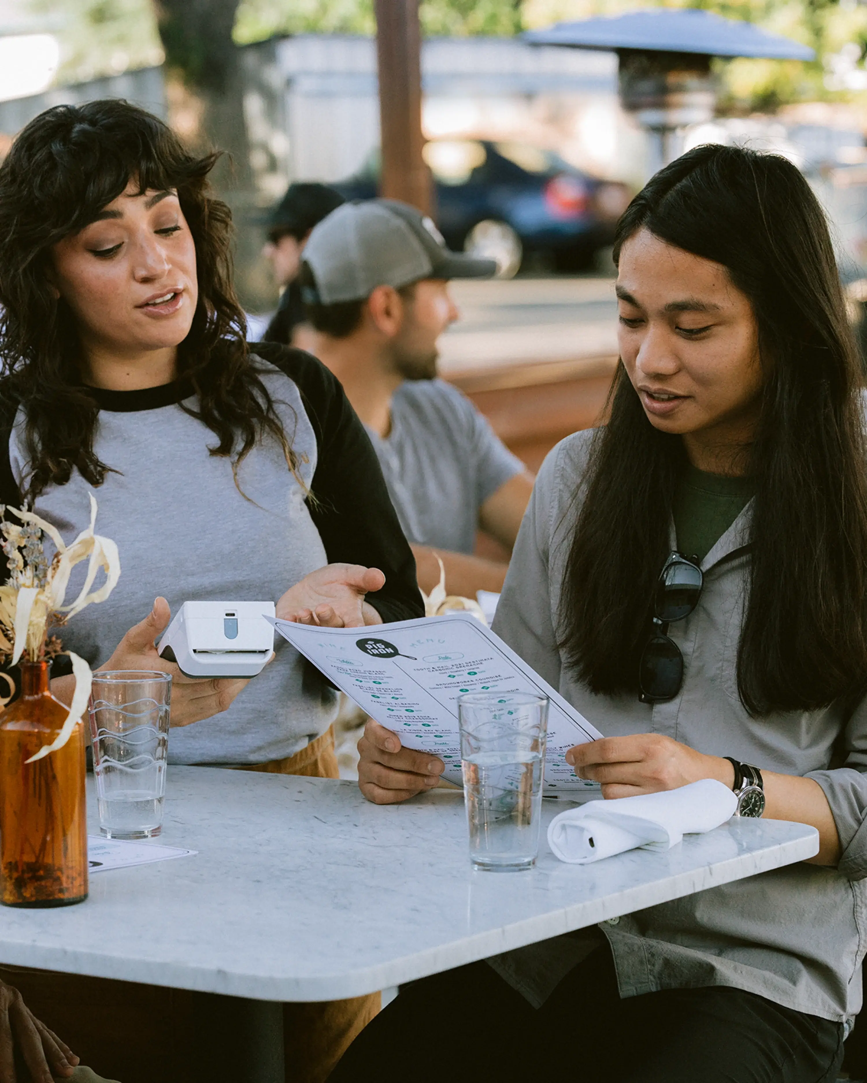 Server holding Clover Flex beside a guest reviewing a paper menu at an outdoor dining table