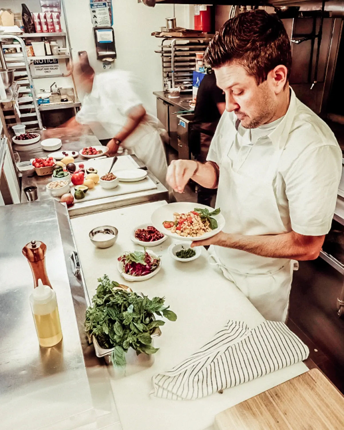 Chef finishing a plated dish in a bright commercial kitchen with stainless prep surfaces
